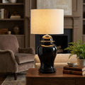 Black ceramic ginger jar table lamp with a cream drum shade on a wooden table in a classic living room setting.
