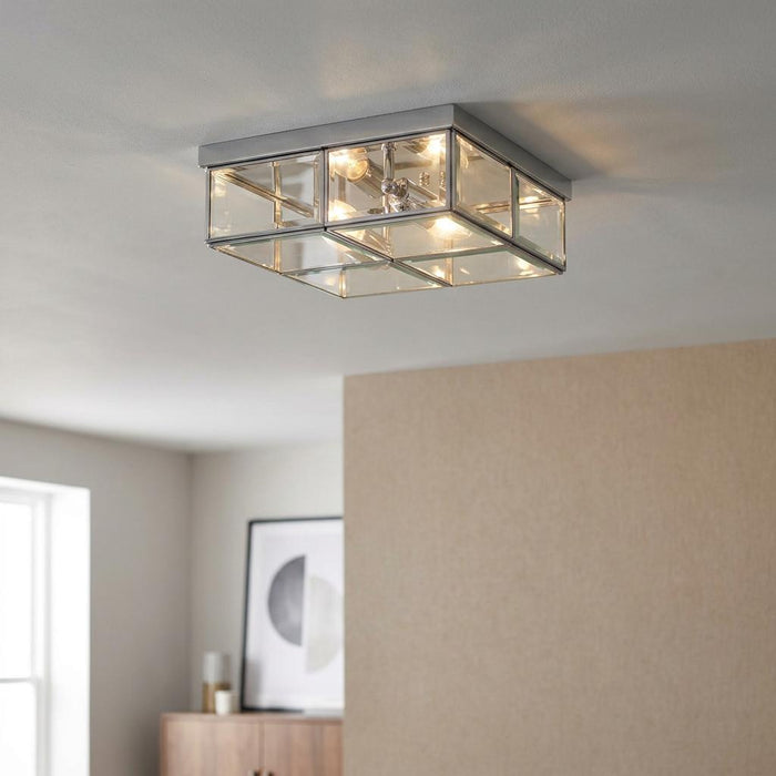 Square chrome and bevelled glass flush ceiling light with glowing bulbs mounted on a white ceiling.