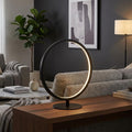 A minimalist black circular LED table lamp illuminates a modern living room from a wooden console table.