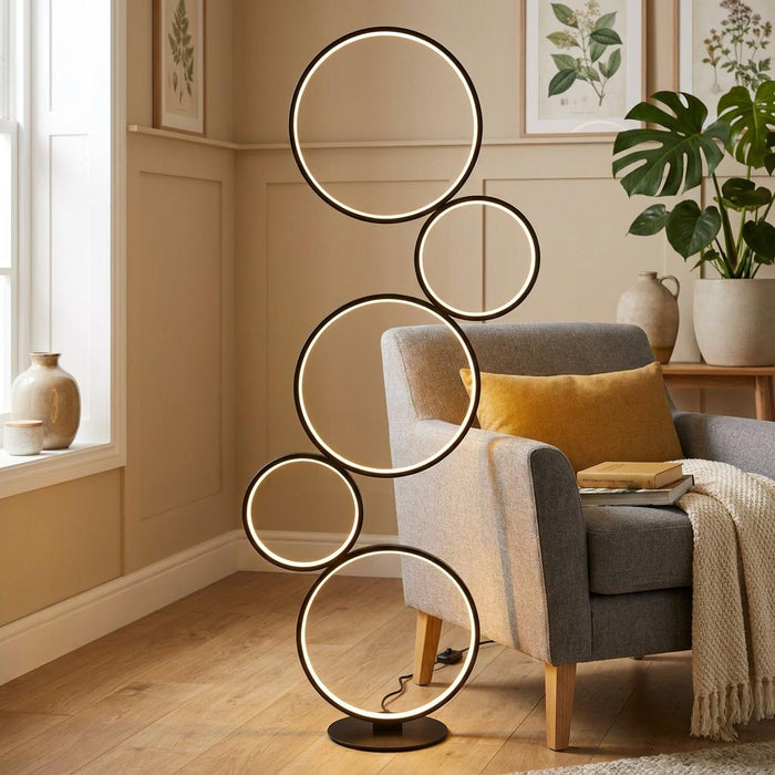 Modern black floor lamp with five glowing LED rings standing in a living room next to a grey armchair.