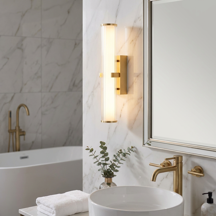 Modern gold bathroom wall light with cylindrical ribbed glass glowing on a marble wall next to a mirror.