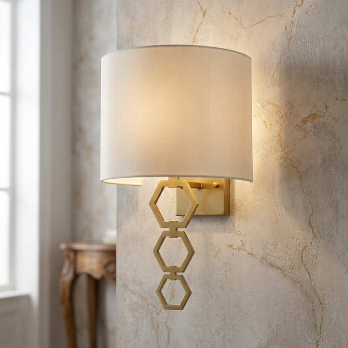 Aged brass wall light with a white drum shade and hexagonal geometric stem on a textured marble wall.