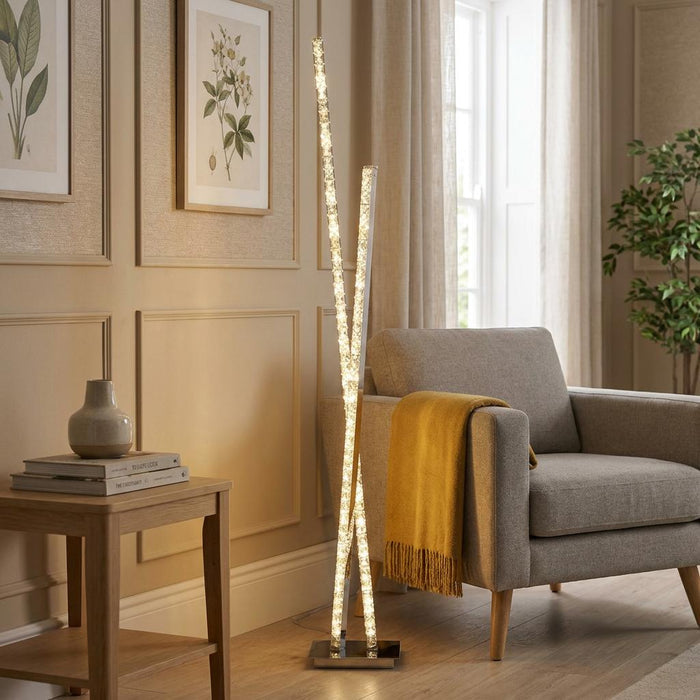 Modern LED floor lamp with two crossed crystal bars stands beside a gray armchair in a living room.