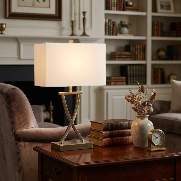 Antique brass table lamp with an X-shaped base and white rectangular shade on a wooden side table in a cozy room.
