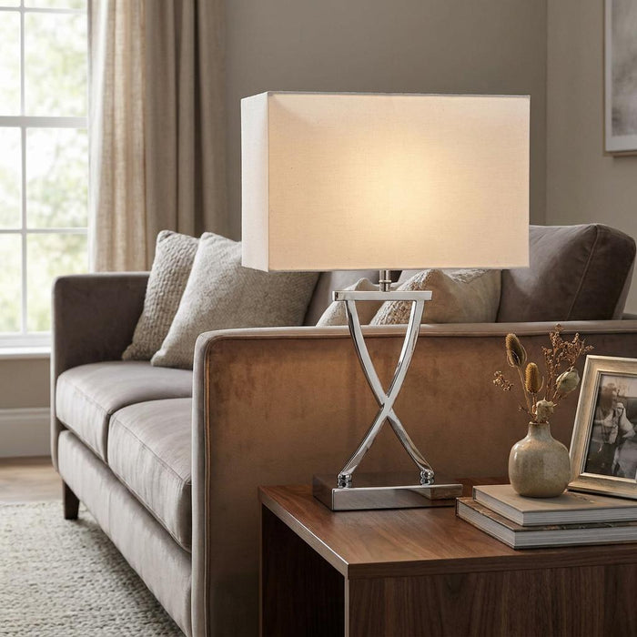Chrome table lamp with a crossed base and white rectangular shade on a side table next to a velvet sofa.