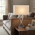 Chrome table lamp with a crossed base and white rectangular shade on a side table next to a velvet sofa.