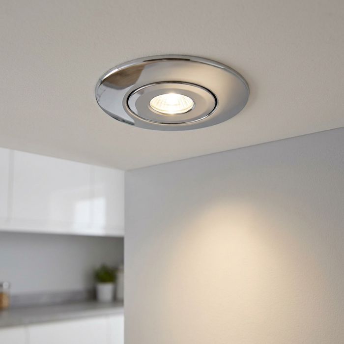Chrome circular recessed spotlight illuminated in a white ceiling, casting light onto a grey wall.