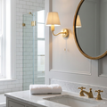 Brass wall light with a beige fabric shade mounted next to a round mirror in a traditional bathroom.