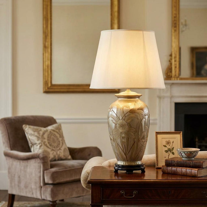 A silver floral ceramic table lamp with a tall ivory empire shade on a wooden side table in a warm living room.