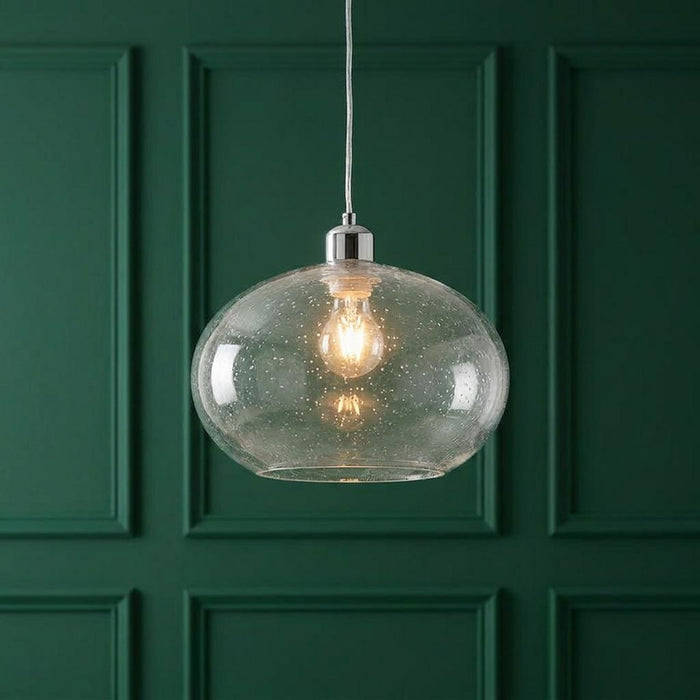 Seeded clear glass pendant light with an oval shade and chrome top, illuminated against a green wood-paneled wall.