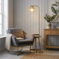 Dimple Modern Brass Floor Lamp with an amber glass globe shade standing next to a grey armchair in a cozy living room.