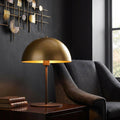 Dome bronze table lamp with a gold metal shade on a dark wooden table beside a grey armchair.