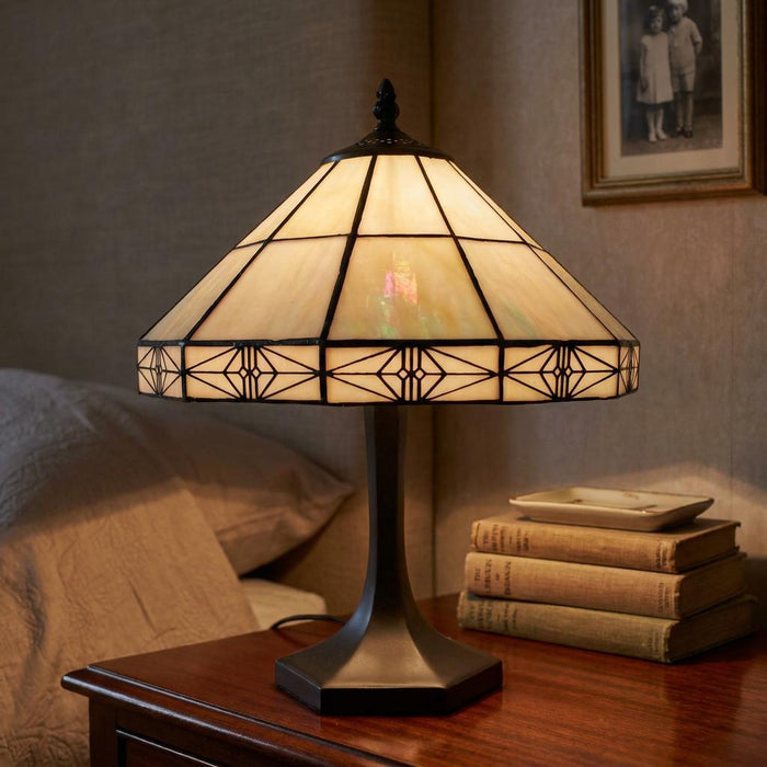 Dorchester Tiffany medium table lamp with cream geometric glass shade on a dark bedside table next to books.