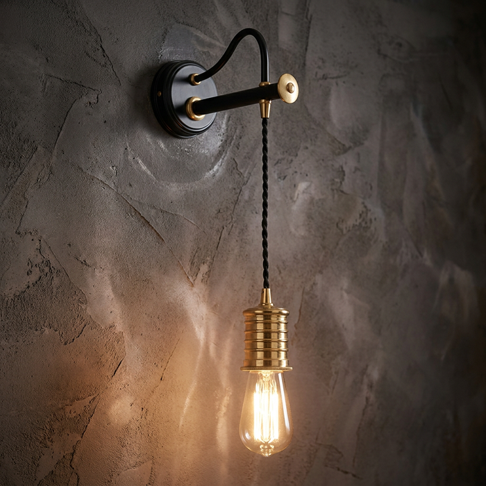Black and brass wall light with a hanging braided cord and glowing Edison bulb against a textured gray wall.