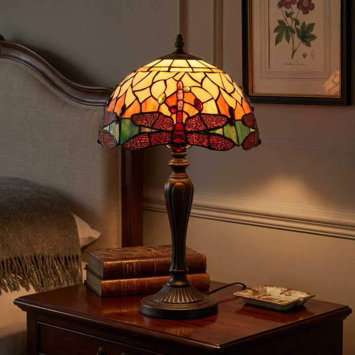 Dragonfly Tiffany table lamp with an orange stained glass shade, lit on a dark wood nightstand in a cozy bedroom.