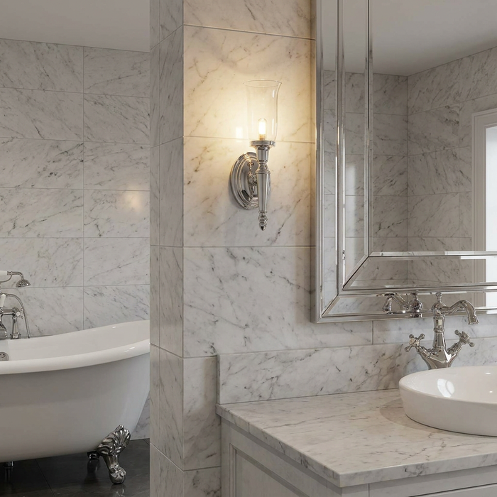 Polished chrome wall light with a clear glass shade on a marble bathroom wall beside a mirror.