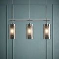 Three-light chrome bar pendant with smokey and frosted glass cylindrical shades against a teal paneled wall.
