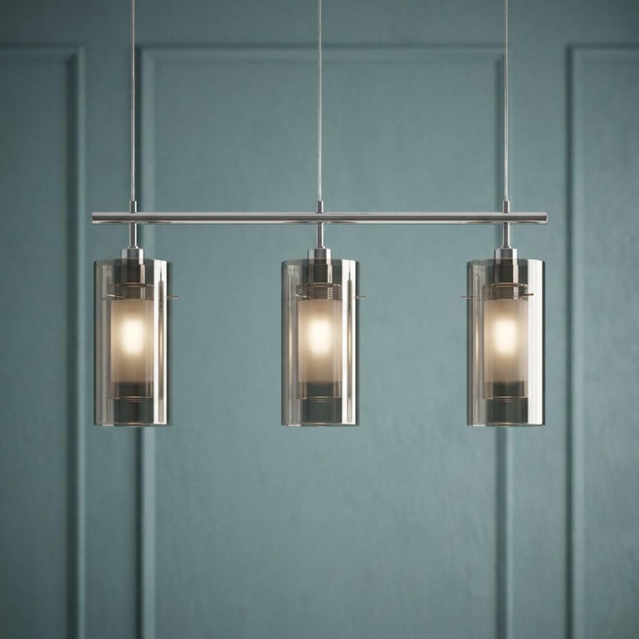 Three-light chrome bar pendant with smokey and frosted glass cylindrical shades against a teal paneled wall.