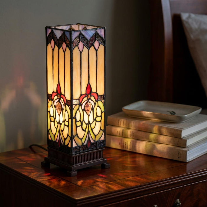 Illuminated Durham square Tiffany table lamp with floral stained glass on a wooden bedside table beside books.