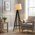 Dark wooden tripod floor lamp with a linen drum shade in a modern living room next to a grey armchair.