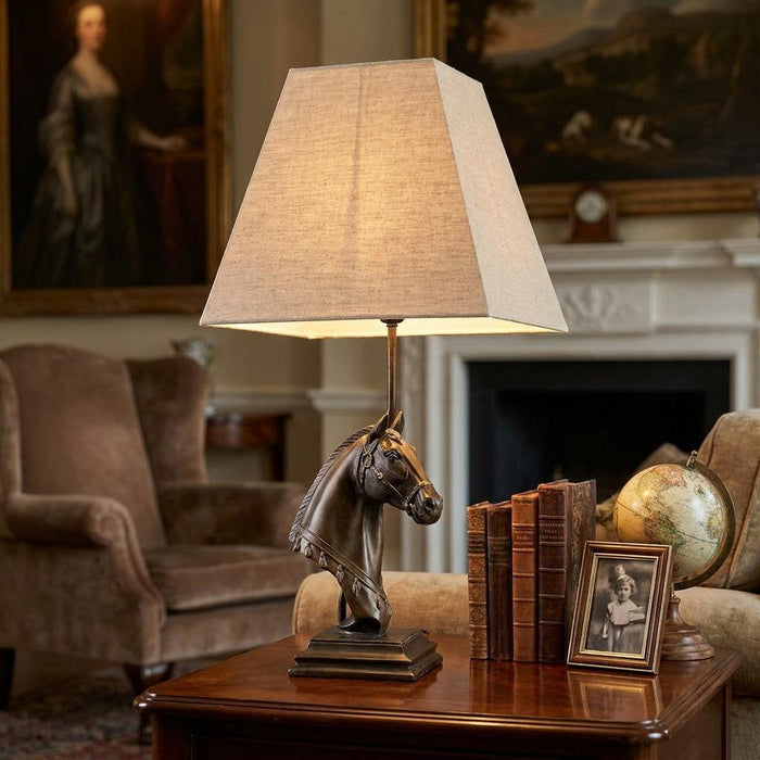 Bronze horse head table lamp with a beige tapered square shade on a wooden table in a traditional living room.