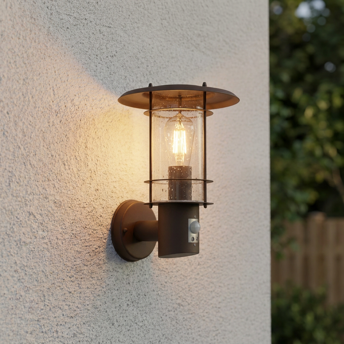 Rust brown lantern wall light with a PIR sensor illuminated on a textured white exterior wall.