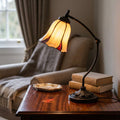 Lit Edinburgh Tiffany swan neck lamp with an amber tulip shade on a wooden table beside books in a cozy room.