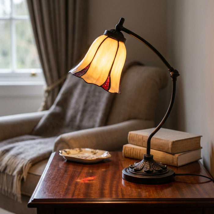 Lit Edinburgh Tiffany swan neck lamp with an amber tulip shade on a wooden table beside books in a cozy room.
