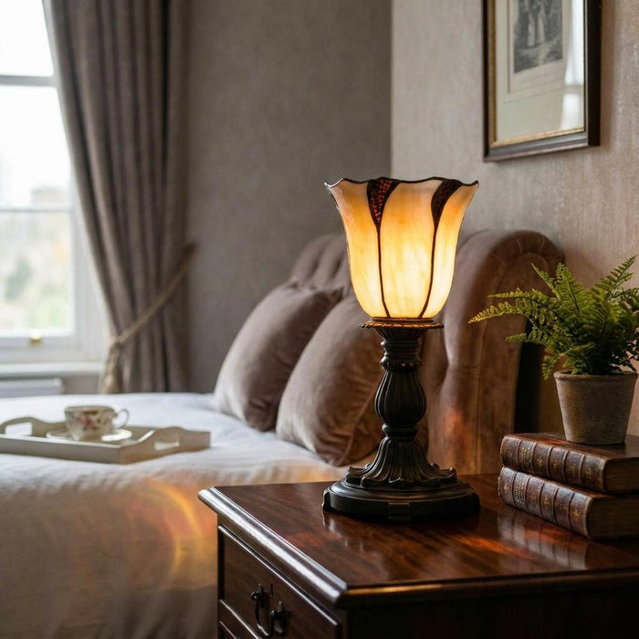 Edinburgh Torchiere Tiffany Bedside Lamp with an amber tulip glass shade glowing on a dark wood nightstand.