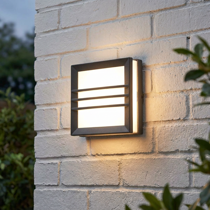 Square dark grey outdoor wall light with horizontal bars emitting warm light on a white brick wall.