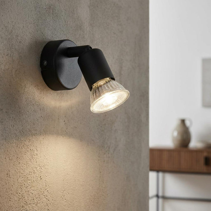 Black wall spotlight with a ribbed glass shade mounted on a textured concrete wall, emitting a warm glow.