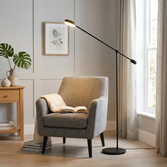 Modern black boom floor lamp illuminating a grey armchair and book in a neutral living room.