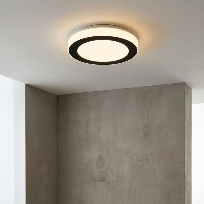 A modern round flush ceiling light with a black ring detail, illuminated against a white ceiling and grey wall.