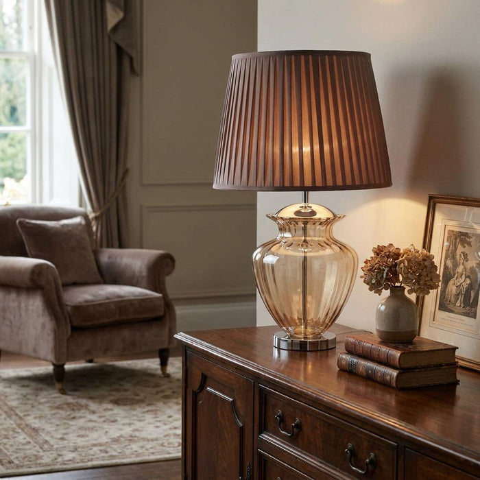 Amber glass urn table lamp with a brown pleated shade on a dark wood sideboard in a cozy living room.