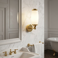 Polished brass wall light with a white glass shade mounted on a marble wall in a traditional bathroom.