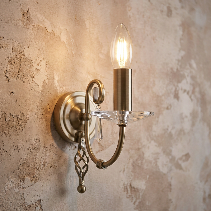 Single-light aged brass wall sconce with crystal droplets and candle bulb glowing against a textured wall.