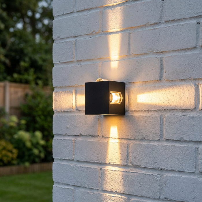 Black cubic outdoor wall light emitting four cross-shaped light beams on a white brick wall.