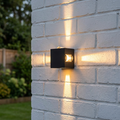 Black cubic outdoor wall light emitting four cross-shaped light beams on a white brick wall.