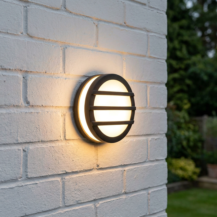 A circular black outdoor wall light with horizontal grilles glows warmly on a white brick wall.