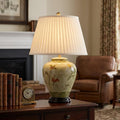 Elstead Arum Lily floral ceramic table lamp with a pleated white shade on a wooden desk in a living room setting.