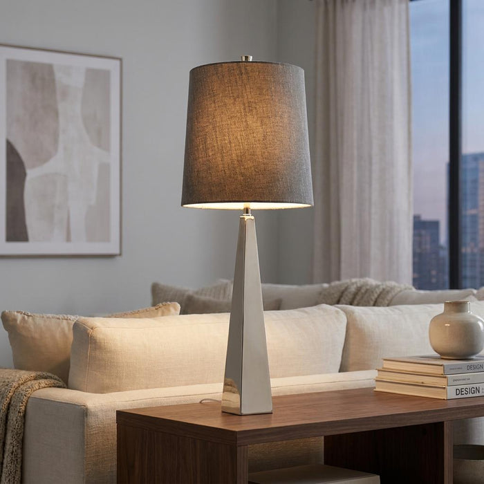 Polished nickel table lamp with a grey fabric shade glows on a wooden table in a modern living room.