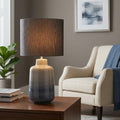Blue and grey ombre ceramic table lamp with a dark drum shade glowing on a wooden coffee table.