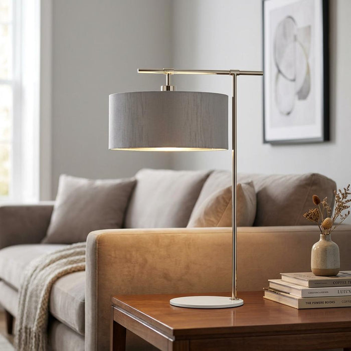 Modern table lamp with a grey drum shade and nickel arm on a wooden side table in a bright living room.