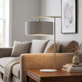 Modern table lamp with a grey drum shade and nickel arm on a wooden side table in a bright living room.