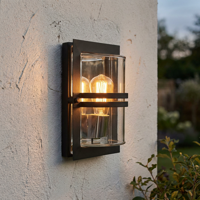 Black outdoor wall light with clear glass and warm Edison bulbs mounted on a white textured wall.