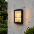 Illuminated Elstead Basel black wall light with frosted glass mounted on a weathered white outdoor wall.