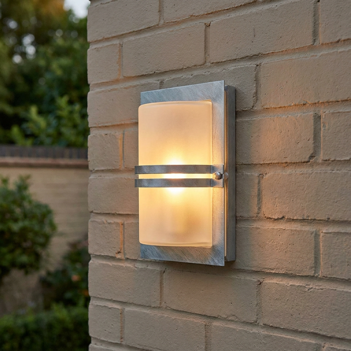 Galvanised steel wall light with frosted glass and a warm glow, mounted on a tan brick exterior wall.