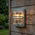 Galvanised steel outdoor wall light with a glowing bulb mounted on a rustic wooden plank wall.