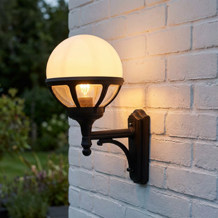 A glowing Elstead Bologna black outdoor uplighter wall light mounted on a white brick wall in a garden.