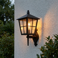 Traditional black lantern wall light with textured glass panes glowing warmly on a white exterior wall.